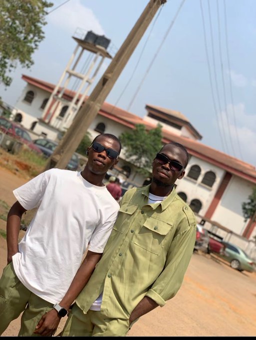Two young men wearing sunglasses pose outdoors in front of a building with red trim, one in a white t-shirt and the other in a green button-up shirt.