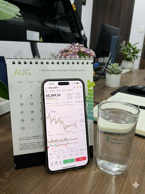A desk scene shows a smartphone displaying Bitcoin (BTC/USDT) trading data at $63,359.25, positioned next to a Northern GeoGrid glass of water and an August spiral calendar.