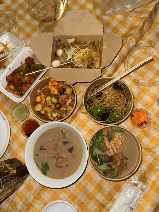 An overhead photo of a dinner spread on a yellow and white checkered tablecloth, featuring bowls of ramen, noodles, fried items, soups, and small sauce dishes with chopsticks.