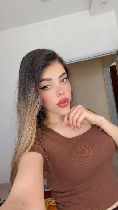 A selfie of a young woman with long brown hair, winged eyeliner, and pink lips, wearing a fitted brown ribbed top.