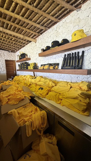 A storage room with a thatched roof and stone walls contains numerous piles of yellow clothing and branded merchandise, with hats and packaged items displayed on wooden shelves.