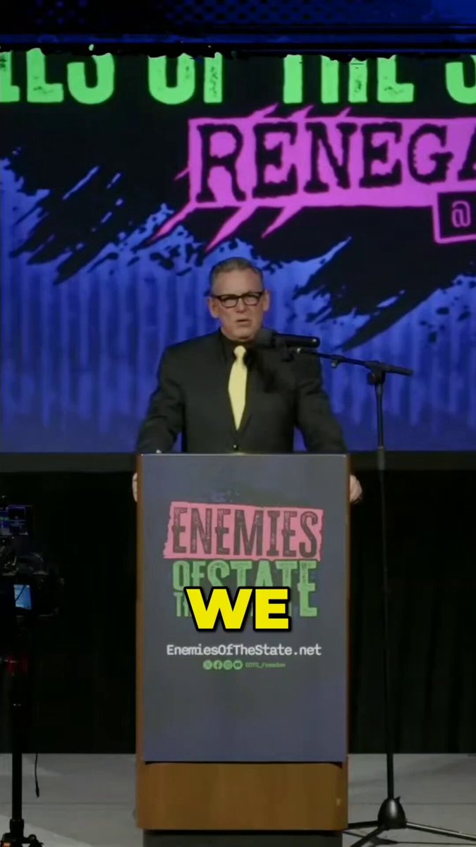 The image features a man standing behind a podium, giving a speech or presentation at an event. The man appears to be wearing glasses and a suit, suggesting that he holds a professional position. In front of him, there's a sign with the words "Enemies of the State" written on it in yellow letters. This sign is likely related to the topic of his speech or presentation.