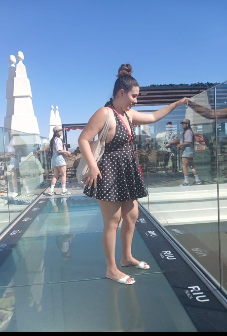 A woman in a polka dot dress and white sandals poses on a glass skywalk at what appears to be the SkyPod observation deck, marked with 'niu' branding.