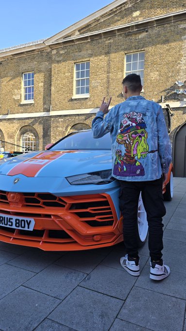 A man wearing a blue denim jacket with a picture of a person on it, standing next to an orange and white car that has a license plate reading "GMS GM Wen Lambo?".