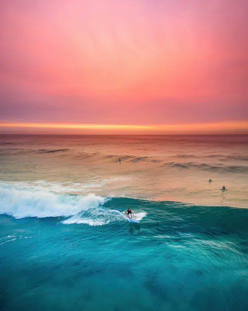 An aerial view of a surfer riding a turquoise wave, with other surfers in the background under a pink and orange sunset sky.