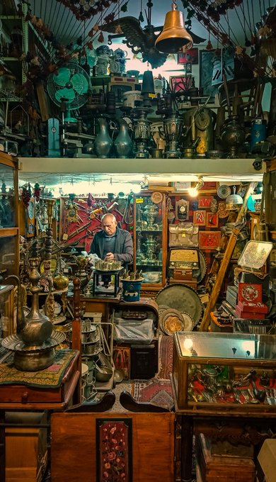 A richly detailed photograph of a man standing inside a densely packed antique shop filled with brass items, clocks, lamps, rugs, and various collectibles.