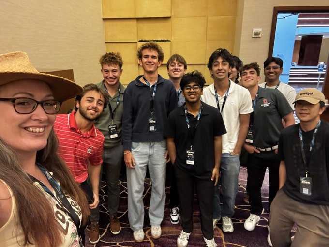 A smiling woman in a straw hat takes a selfie with a group of young men wearing lanyards in a hotel conference room.