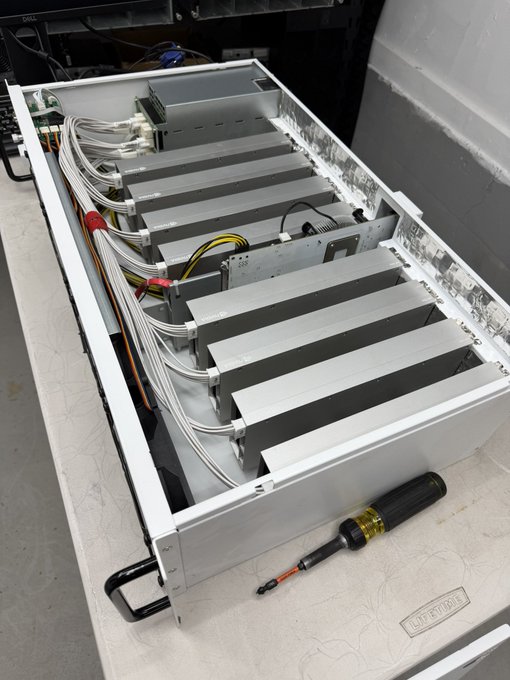 Photograph of an open server/mining rig chassis containing multiple gray mining units connected by cables, with a yellow and black screwdriver resting on the table in front.