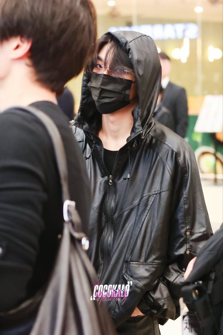 A person wearing a black hooded jacket and black face mask stands in what appears to be an airport terminal, with another individual carrying a bag partially visible in the foreground. The watermark "COCKALO" appears at the bottom, matching the airport fashion style often featured in the "261019" dated post about Xinlong from AlphaDriveOne.