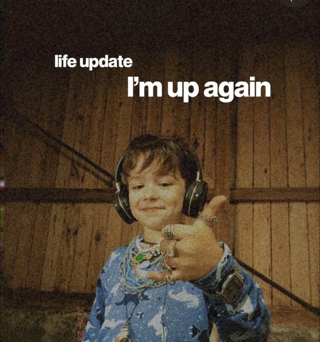 A vintage-style photograph of a young child wearing large headphones and a blue patterned shirt giving a thumbs-up, with overlay text reading 'life update' and 'I'm up again.'