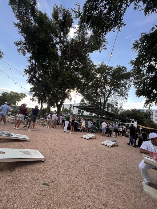 Outdoor gathering at a venue with large trees, string lights, cornhole boards scattered on the gravel, and groups of people socializing in the early evening.