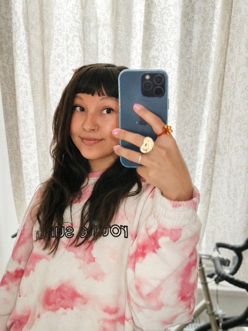 A young woman with long dark hair takes a mirror selfie with her blue smartphone, wearing a pink tie-dye 'You're So' sweatshirt.
