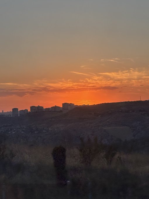 A scenic sunset view over a hilly landscape with city buildings on the horizon, silhouetted against an orange and pink sky.