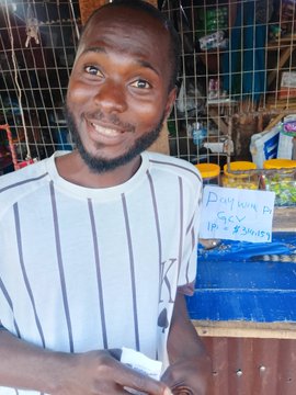 A smiling young man stands inside a small shop, holding what appears to be purchased goods, with a handwritten sign reading "PAY WITH $Pi" and "1pc 18 = ₦3500" visible behind him, illustrating the post's claim of a Pi Network vendor accepting the cryptocurrency for food items in Africa.