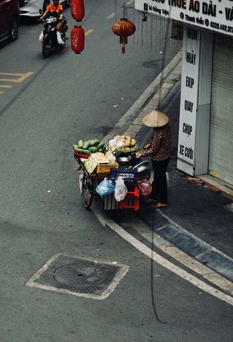 An overhead view shows a street vendor wearing a conical hat standing next to a cart filled with fruits and vegetables on a sidewalk in front of a building with red lanterns hanging above. Text in Vietnamese is visible on the building's signage.