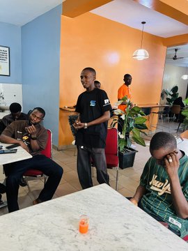 A casual photo of people in a café or restaurant with orange walls, featuring a man standing in the center wearing a dark shirt.