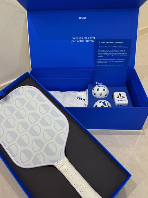 A blue Trust Wallet 9th anniversary merchandise box displays a white pickleball paddle with a Trust logo pattern, along with branded pickleballs and a card reading "Thank you for being part of the journey" and "9 Years, On Your Own Terms," celebrating the crypto wallet's milestone anniversary.