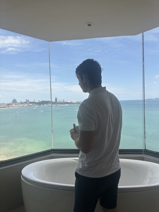 A silhouetted man in a white polo shirt stands beside a round white bathtub, looking out through large angular floor-to-ceiling windows at a turquoise ocean and distant city skyline.