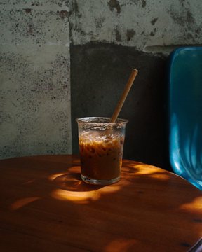 A glass of iced coffee with a straw sits on a wooden table next to a blue chair, with dramatic sunlight casting shadows across the scene.