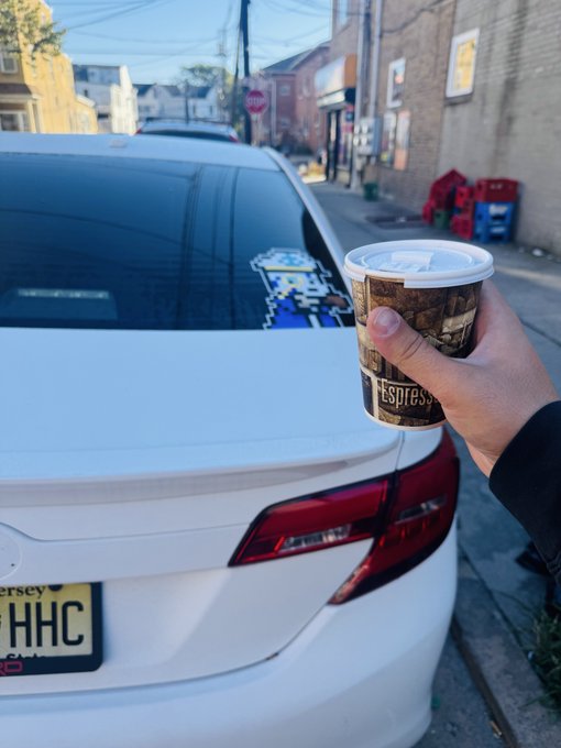 A hand holds an Espresso coffee cup in front of a white Volkswagen with New Jersey license plate 'HHC', parked on a street.
