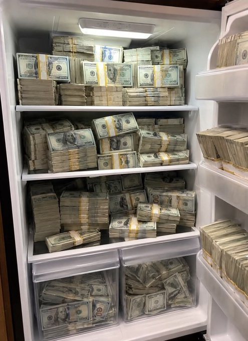 An open refrigerator completely packed with stacks of US hundred-dollar bills arranged in bundles on the shelves and in the drawers.