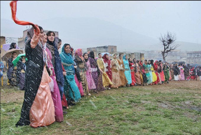 A line of women in colorful traditional dresses holding hands while dancing in a line on a grassy field during a snowfall, with mountains visible in the background.