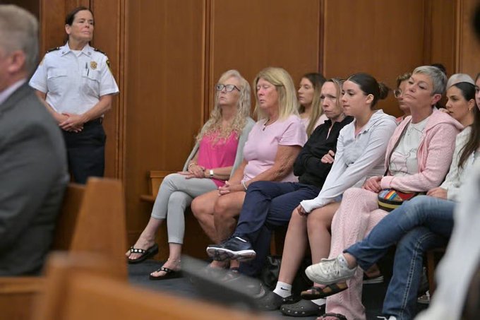 A courtroom scene with several women seated in the gallery, a uniformed officer standing against the wood-paneled wall, and a man in a suit partially visible in the foreground.