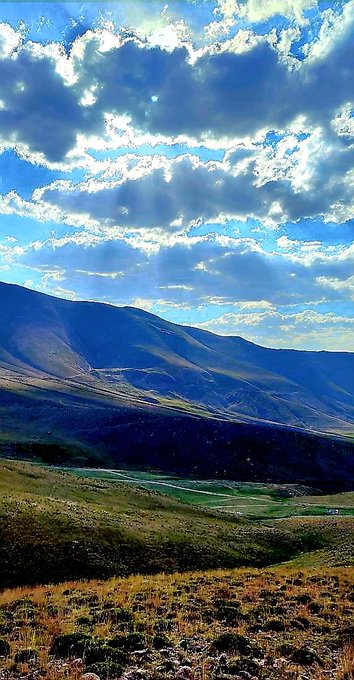 A vertical landscape photo showing a vast mountain valley with green and brown grassy terrain under a bright blue sky with scattered clouds and visible sun rays.