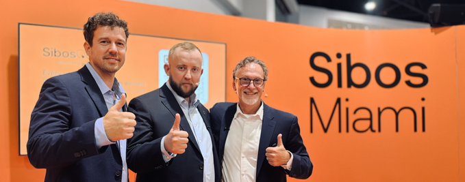 Three men in business attire giving thumbs up while standing in front of an orange wall displaying 'Sibos Miami' in large black letters.