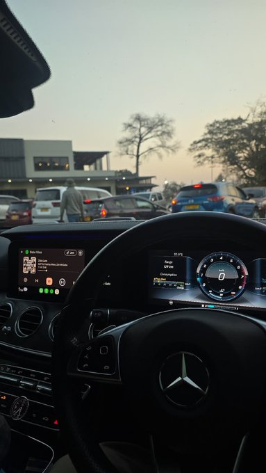 A view from inside a Mercedes-Benz showing the dashboard displays, including a music player and digital instrument cluster showing 'Range: 107 km' and 'CONS: 3.0', with a traffic scene visible through the windshield at dusk.