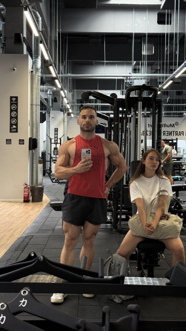 A muscular man in a red tank top takes a mirror selfie at a gym while a woman in a white t-shirt sits nearby on a bench, fitting the "average STONK holder" joke by contrasting his elite physique with a casual gym-goer in the background.