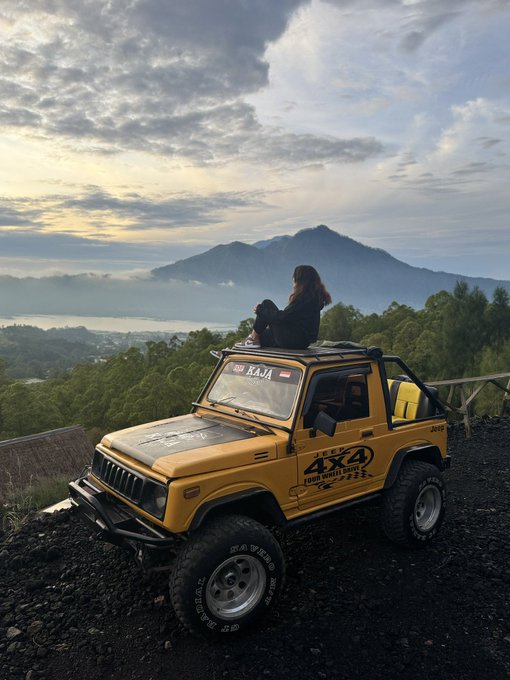 A woman sits atop a yellow 4x4 Jeep (marked "KA" and "4X4 ONE WHEEL DRIVE") overlooking a misty mountain landscape at sunrise, suggesting a scenic getaway. The caption "GM Frens happy moonpay fridayy you all got the $1k last night right?" appears unrelated to the image, as it references a cryptocurrency payment notification rather than the peaceful outdoor scene depicted.