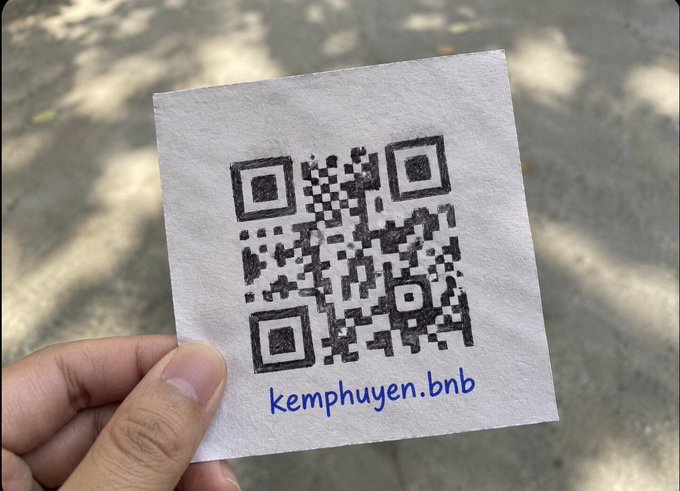A hand holds a small piece of paper printed with a QR code and the text "kemphuyen.bnb," suggesting a crypto payment address on the BNB Smart Chain. The image relates to the post's theme of crypto risks by implying that despite all the dangers in DeFi—rugs, hacks, liquidations, scams, dumps, depegs, and suspicious signatures—you can still safely transact using this QR code.