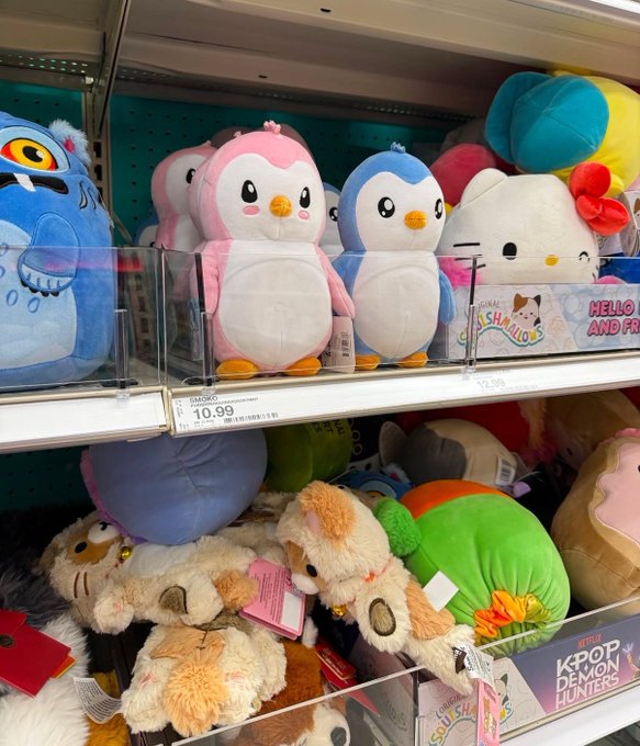 A retail store shelf displaying plush toys including Hello Kitty, penguin characters, and various animals, with a visible price tag of $10.99 and a 'KPOP DEMON HUNTERS' label.