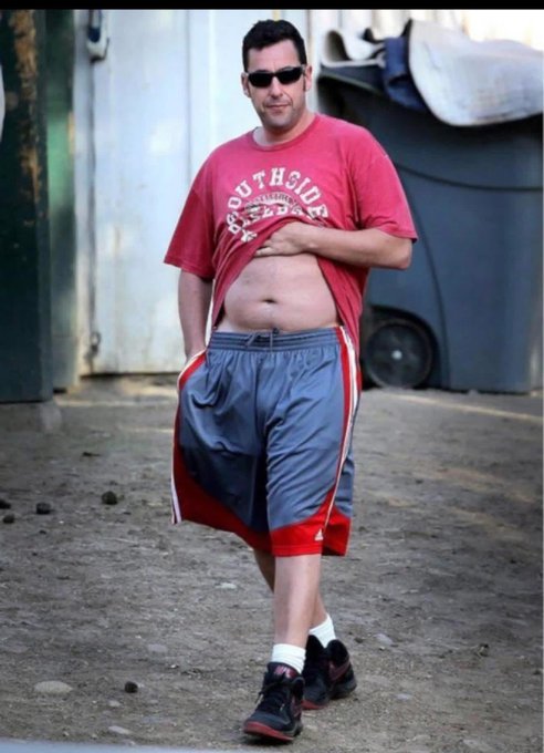 Adam Sandler in a red 'Southside' t-shirt, grey athletic shorts, and sunglasses walking outdoors while lifting his shirt to expose his stomach.