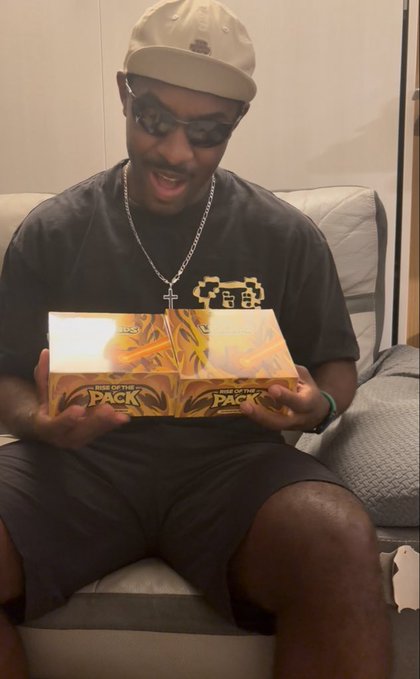 A man wearing a white cap, sunglasses, and a black t-shirt with a pixel art logo holds up two boxes of trading cards labeled 'RISE OF THE PACK,' appearing excited.