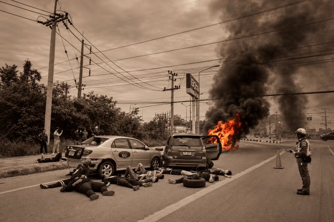 A dramatic scene shows a car engulfed in flames and black smoke on a road, with emergency responders and individuals lying on the ground nearby.