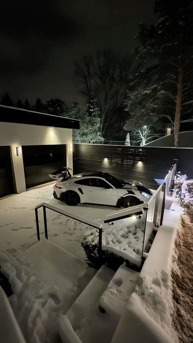 A white Porsche sports car parked on a snow-covered driveway at night, illuminated by outdoor lights next to a modern building.