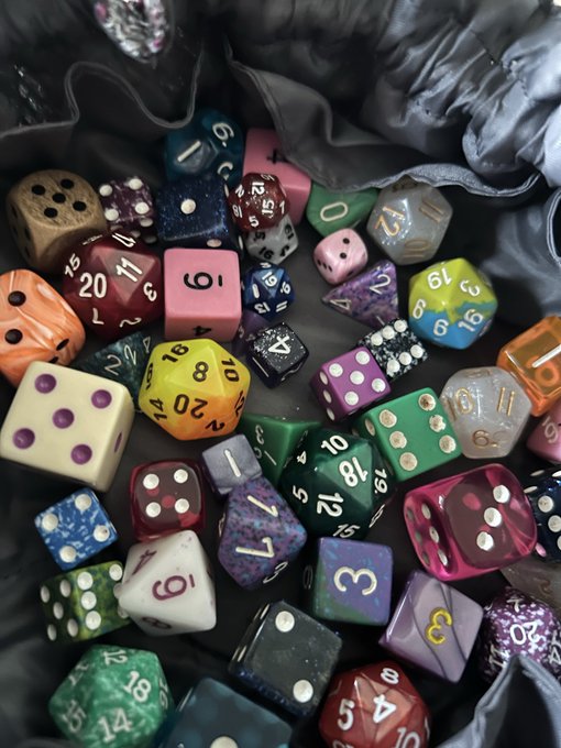 A colorful assortment of polyhedral and other gaming dice scattered inside a black fabric bag, including d20s, d6s, and dice with various colors and patterns like glitter and swirls.