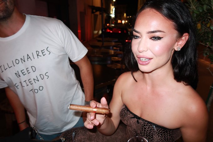 A woman in a brown strapless dress smiles while holding a cigar, standing next to a man wearing a white t-shirt that reads "BILLIONAIRES NEED FRIENDS TOO," capturing a glamorous nightlife scene that mirrors the celebrity-driven, high-roller crypto culture highlighted in the post about the $ANSEM creator coin.