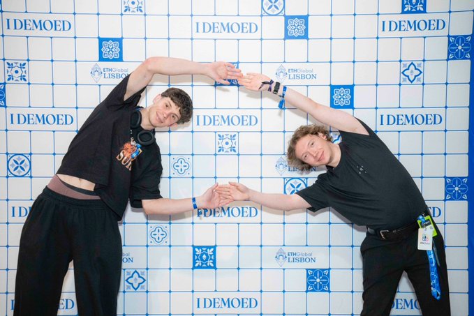 Two young men pose together in front of an "I DEMOED" backdrop at ETHGlobal Lisbon, forming a heart shape with their raised arms as they await the finalist judging results before the closing ceremony.