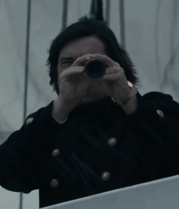 A cinematic shot of a person with dark hair wearing a dark coat with gold buttons, holding a camera up to their face as if taking a photograph.