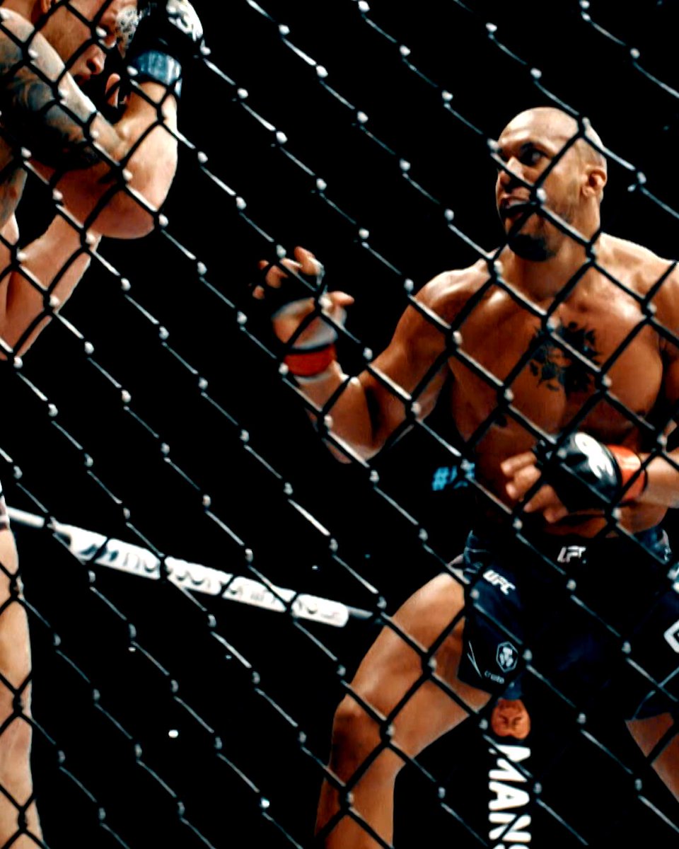 The image shows two men fighting inside a cage, with one man wearing black shorts and the other wearing blue shorts. They are both holding gloves as they engage in combat. The fighter on the left has his head down, while the fighter on the right is looking up at him. The fighters appear to be in the middle of an intense match or practice session.
