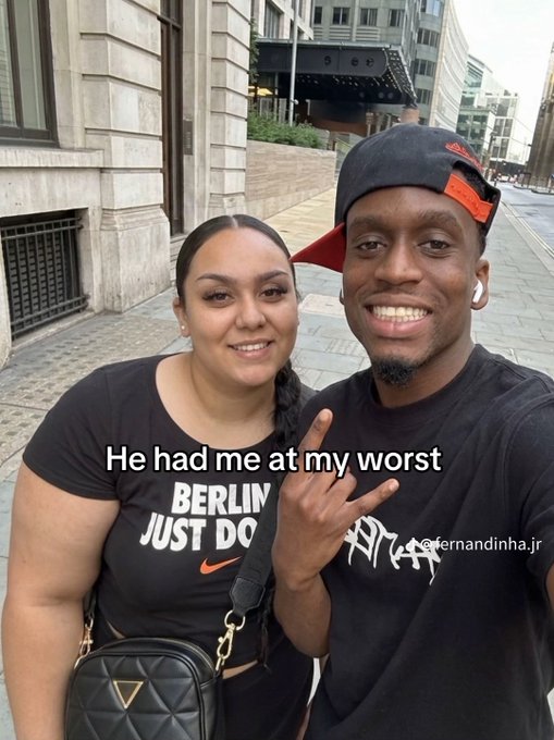 A smiling couple takes a selfie on a city sidewalk; the woman wears a black "BERLIN JUST DO" t-shirt and the man wears a black cap and t-shirt, with the overlaid text "He had me at my worst".
