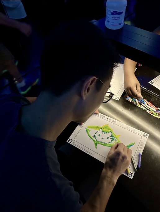 A young man wearing glasses is seen from behind, drawing a green cartoon cat on a 'Crypto Wallet' activity sheet with crayons. A bottle of alcohol wipes is visible on the table nearby.