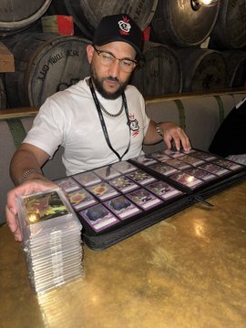 A bearded man wearing glasses and a baseball cap displays rows of dog-themed trading cards from a binder behind stacks of cards on a wooden counter, with whiskey barrels labeled "LINKWOOD" visible in the background. The image ties to the post by showcasing the creator's collection of "Doginal dog" NFTs alongside an appearance of the collector himself.