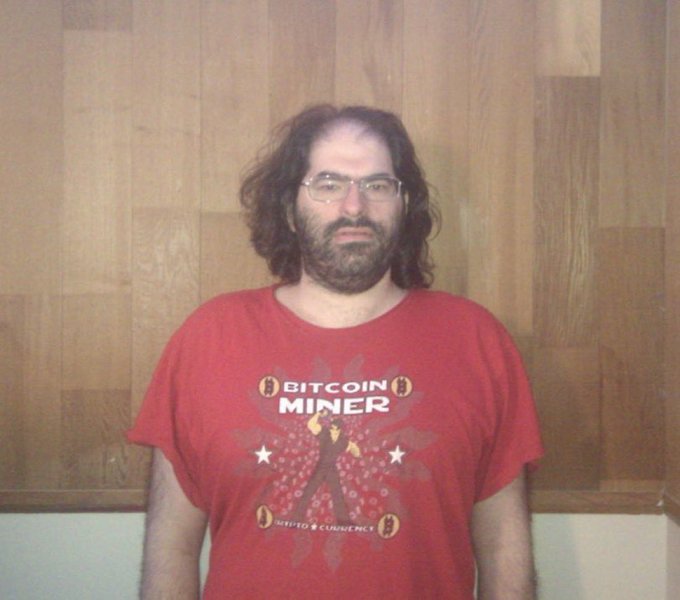 A man with long curly hair, a beard, and glasses stands in front of a wooden paneled wall, wearing a red "BITCOIN MINER - ACCEPTED CURRENCY" t-shirt featuring a design reminiscent of a flag with stars and crossed tools, identified in the post as "The dev."