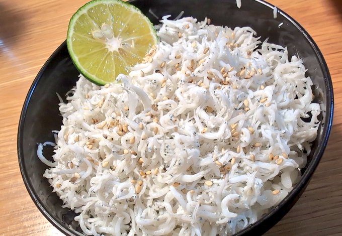 A black bowl filled with whitebait (shirasu) sprinkled with sesame seeds and garnished with a half slice of sudachi (Japanese citrus), served on a wooden table.