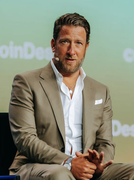 A man with dark hair and a beard wearing a beige suit jacket and white shirt, speaking at what appears to be a CoinDesk conference panel.
