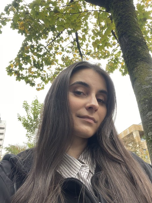 A woman with long dark hair takes a selfie outdoors beneath a tree with green leaves, wearing a striped shirt and black jacket, as she shares the news of her election to the Intersect Product Committee and Growth & Marketing Committee seats for Cardano.
